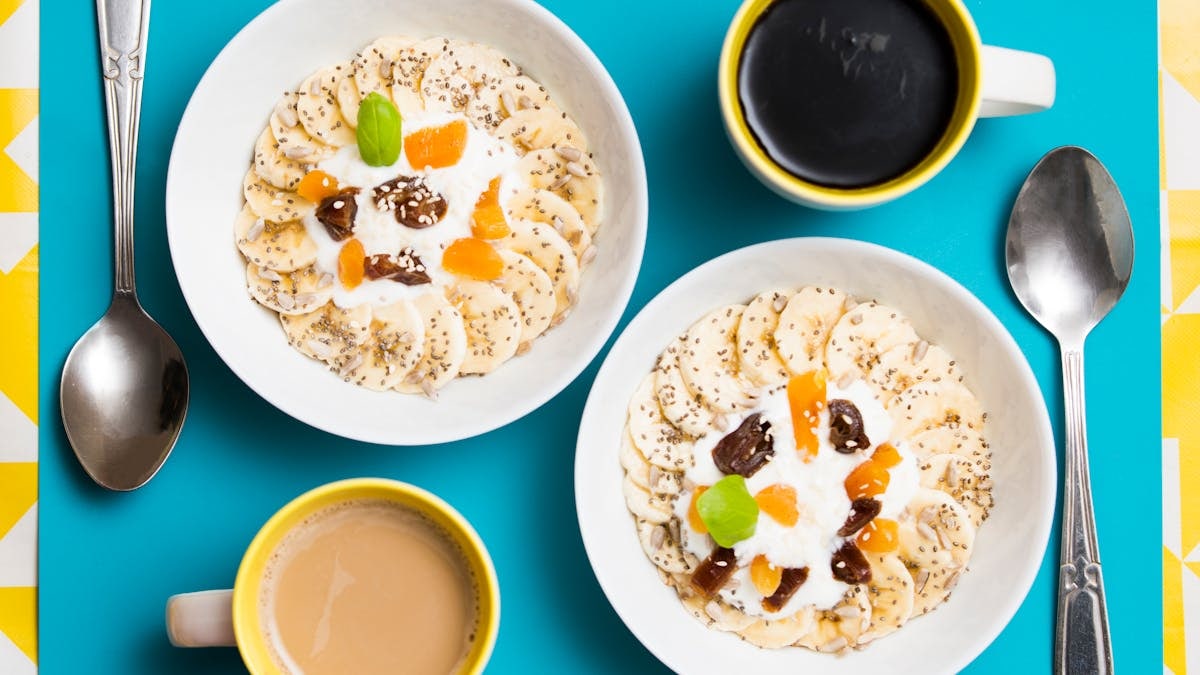 Top view of two breakfast bowls with bananas, yogurt, and coffee cups on a vibrant background.